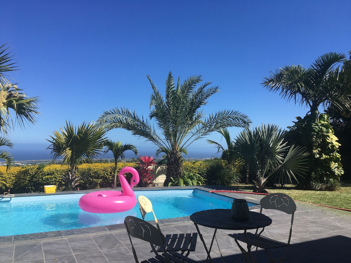 A private pool is surrounded by tropical palm trees and lush greenery. A pink inflatable float is placed in the clear water, and a small seating area with two chairs and a table offers a spot to enjoy the view of the vibrant landscape beyond.