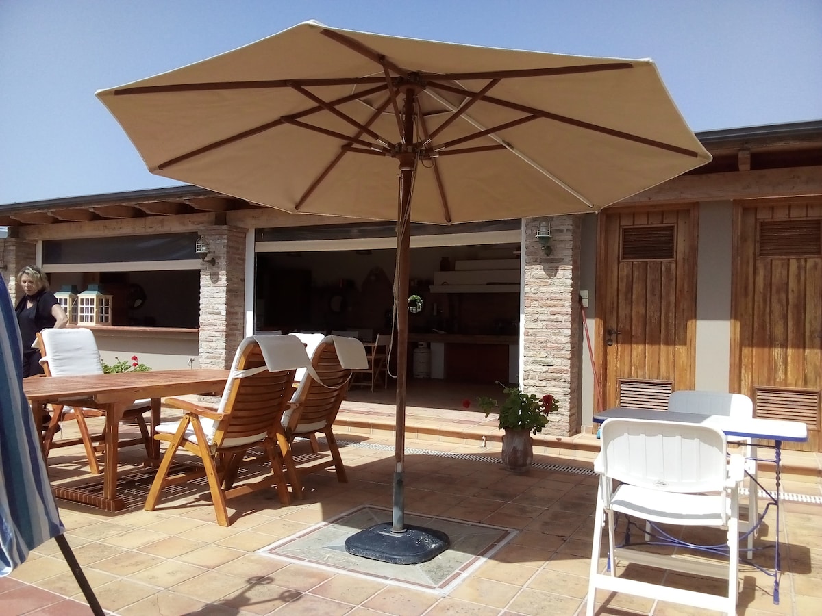 A shaded outdoor dining area is presented, featuring a large umbrella over a wooden table accompanied by several folding chairs. Behind the table, a view of a kitchen area can be seen through open sliding doors. Potted plants add a touch of greenery to the space.