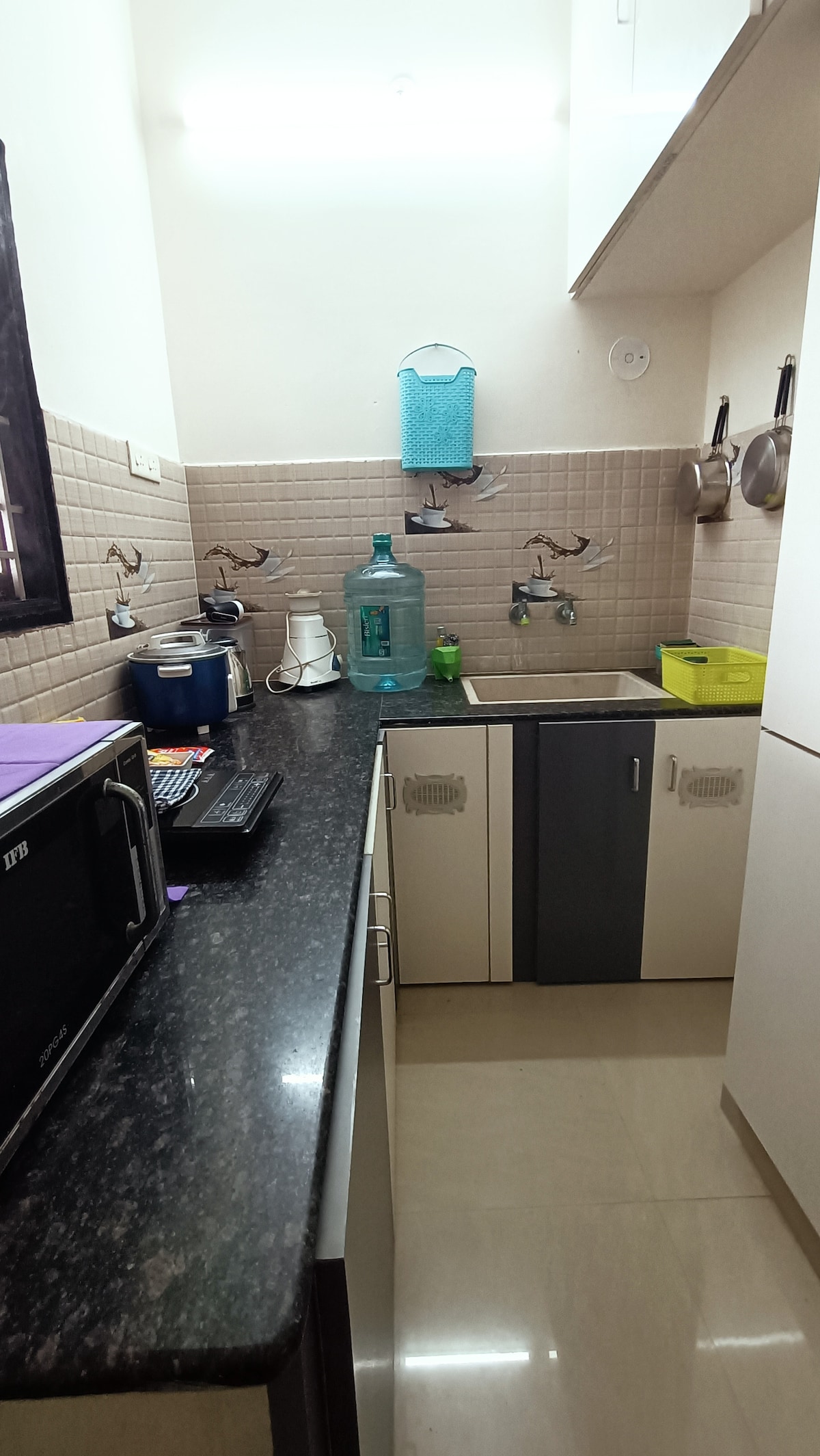 The kitchen space features a sleek, dark countertop complemented by a light tiled backsplash. Essential appliances, including a microwave and a gas stove, are visible. Functional storage is provided by cabinets above and below the counter. A large water dispenser and various utensils are organized for convenience.