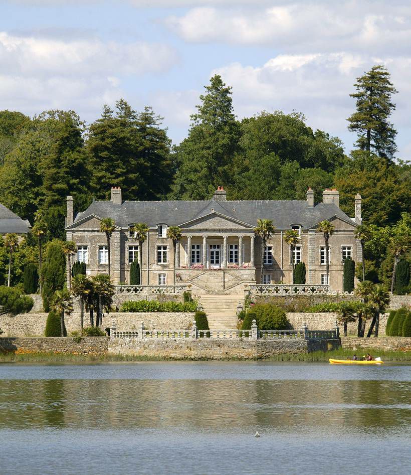 A stately structure is situated by a serene lake, surrounded by lush greenery and tall trees. The building features a symmetrical design with multiple stories, decorative stonework, and a grand entrance. A small boat can be seen gliding on the water, completing the tranquil scene.