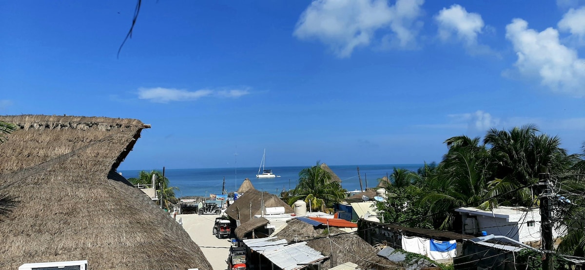 A panoramic view of the coastline features a clear blue sky with scattered clouds, overlooking the ocean. In the foreground, thatched roofs and greenery are seen, while a boat is visible in the water, enhancing the serene coastal atmosphere.
