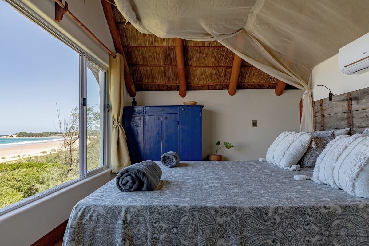 A spacious bedroom features a large double bed adorned with soft textiles and plush pillows. Natural light floods the room through a sizable window, offering a view of the beach and coastal greenery. A blue cabinet adds a pop of color to the earthy tones.