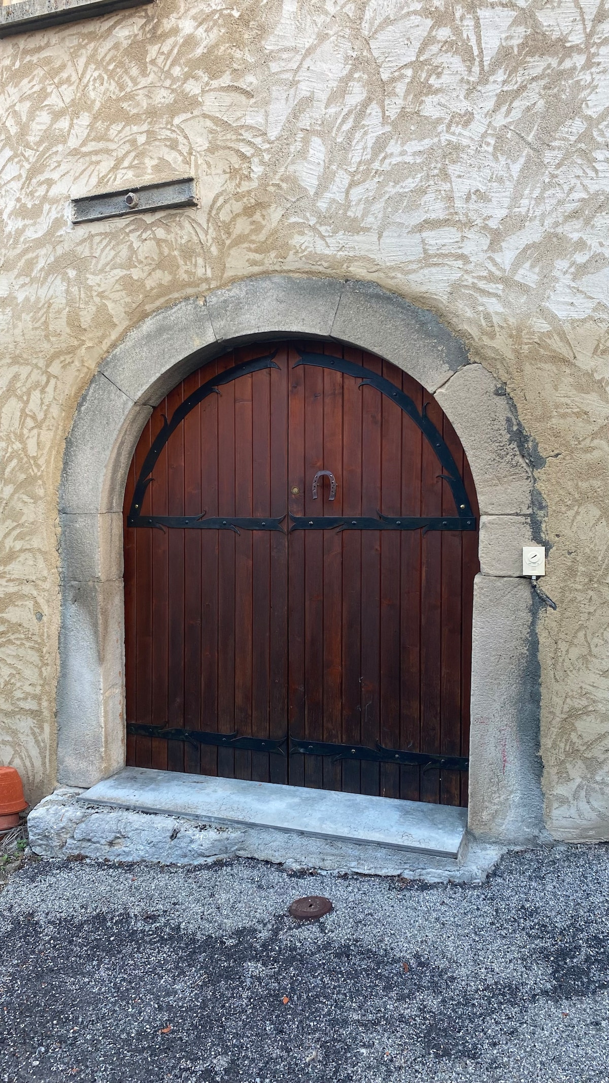 An arched wooden door is framed by a textured stone wall. The door features dark wood panels secured with wrought iron fittings, including a decorative handle. A stone threshold is visible at the base, contrasting with the surrounding gravel surface.