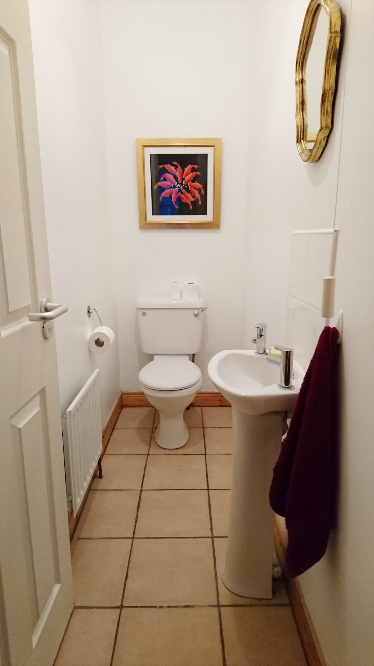 A bathroom is presented with a white toilet and a small sink beside it. Decorative art is hung on the wall above the sink, featuring a colorful floral design. Neutral tiles cover the floor, and a towel is neatly placed on the sink's edge.