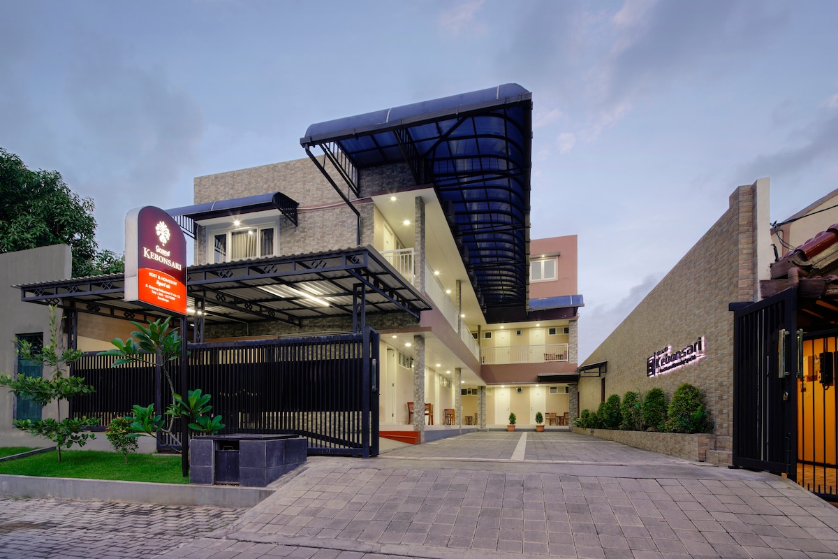 The exterior of the Grand Kebonsari Guesthouse is presented, featuring a modern design with a spacious entrance. A covered walkway leads to the entrance, flanked by greenery. The signage is prominently displayed, and the overall structure is well-lit, showcasing a welcoming atmosphere.