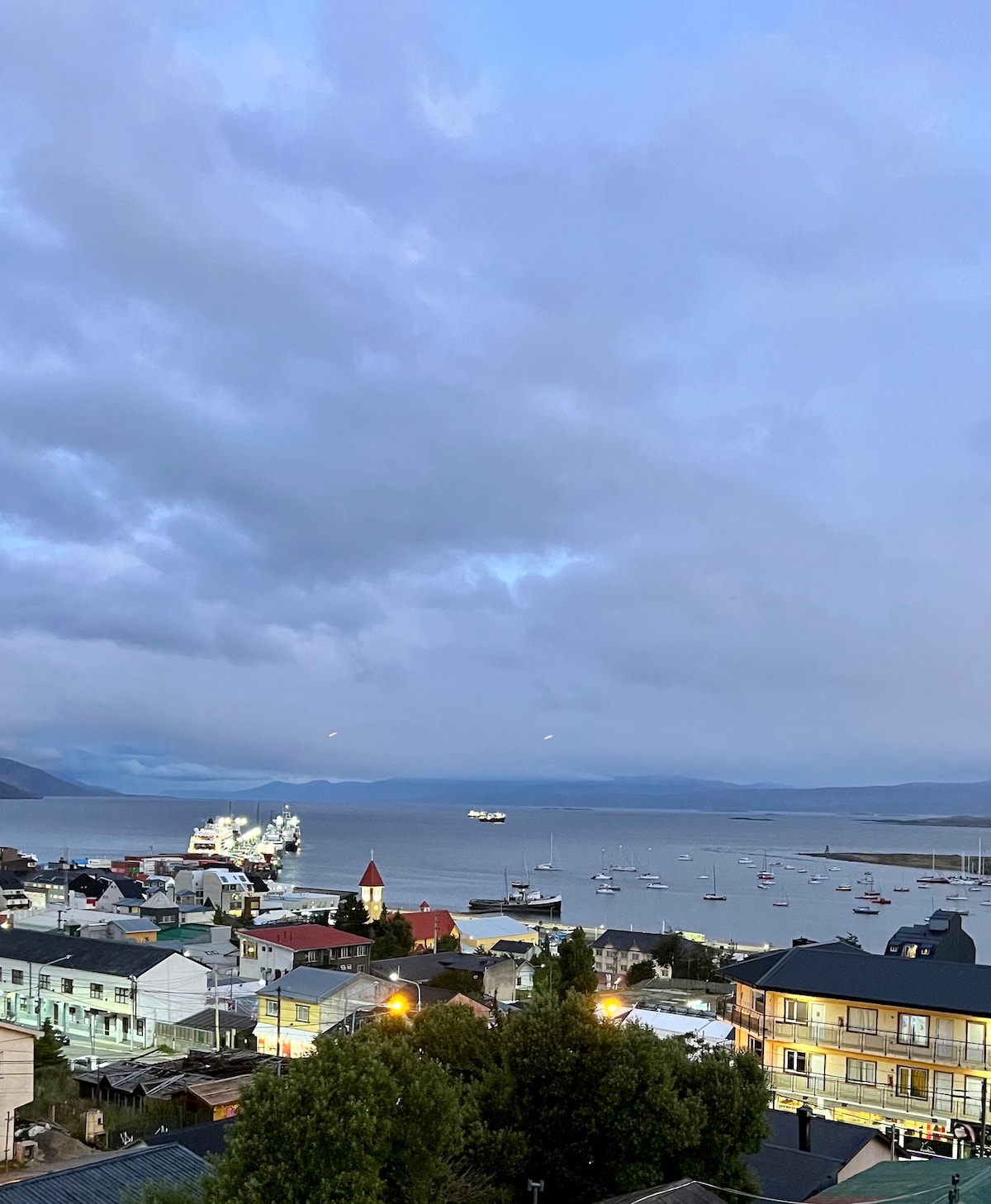 A coastal view reveals a harbor filled with boats, surrounded by quaint buildings. Gentle waves lap against the shore under a cloudy sky, offering a serene atmosphere. The distant mountains provide a subtle backdrop, enhancing the peaceful scenery.