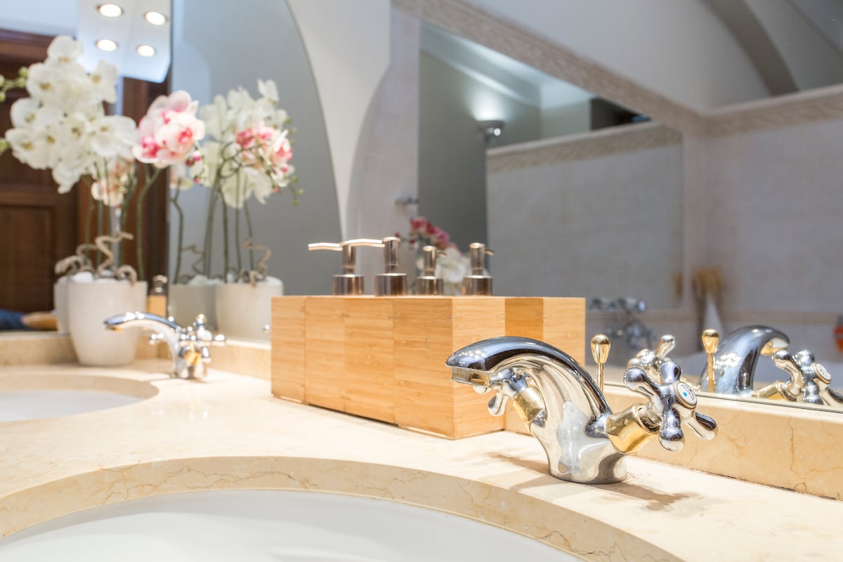 A well-appointed bathroom features elegant chrome faucets positioned above a marble countertop. A wooden organizer holds various toiletries, while soft white orchids are displayed in pots, adding a touch of natural beauty to the space.
