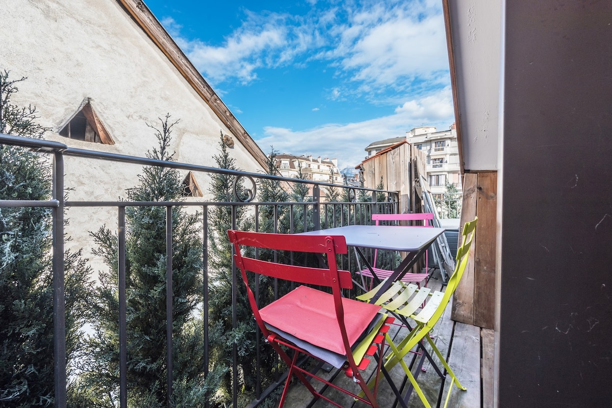 A small balcony features a simple table surrounded by four colorful chairs in pink, green, and yellow. The outdoor space is framed by greenery and offers views of nearby buildings under a clear blue sky.