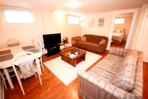 The spacious living area features a comfortable sofa and a casual couch, alongside a dining table set for four. A flat-screen television is positioned on a media unit, while a coffee table sits centrally on a light area rug. Bright windows provide natural light.