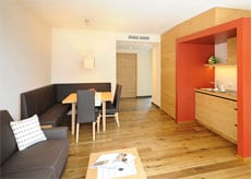 A modern living space is shown, featuring a dining area with a wooden table and several grey chairs. A cozy couch sits in the corner, complemented by a decorative pillow. The kitchen area includes wooden cabinetry and minimalist design elements, highlighted by warm wooden flooring throughout.