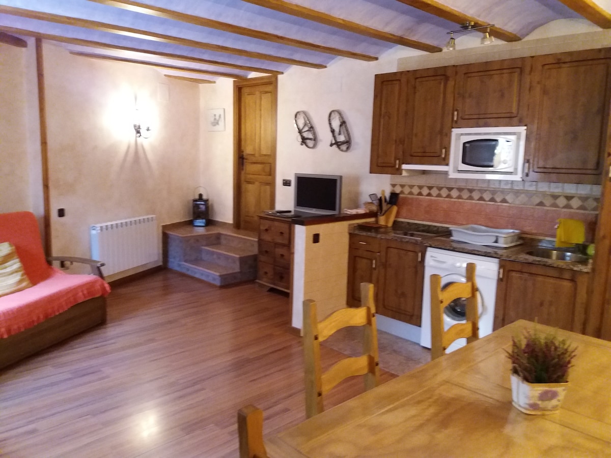 A cozy living space is displayed featuring light wood beams and warm wall tones. A sofa bed sits beside a rustic dining table with wooden chairs. A kitchen area with wooden cabinets includes modern appliances. Natural light fills the room from a wall-mounted window.