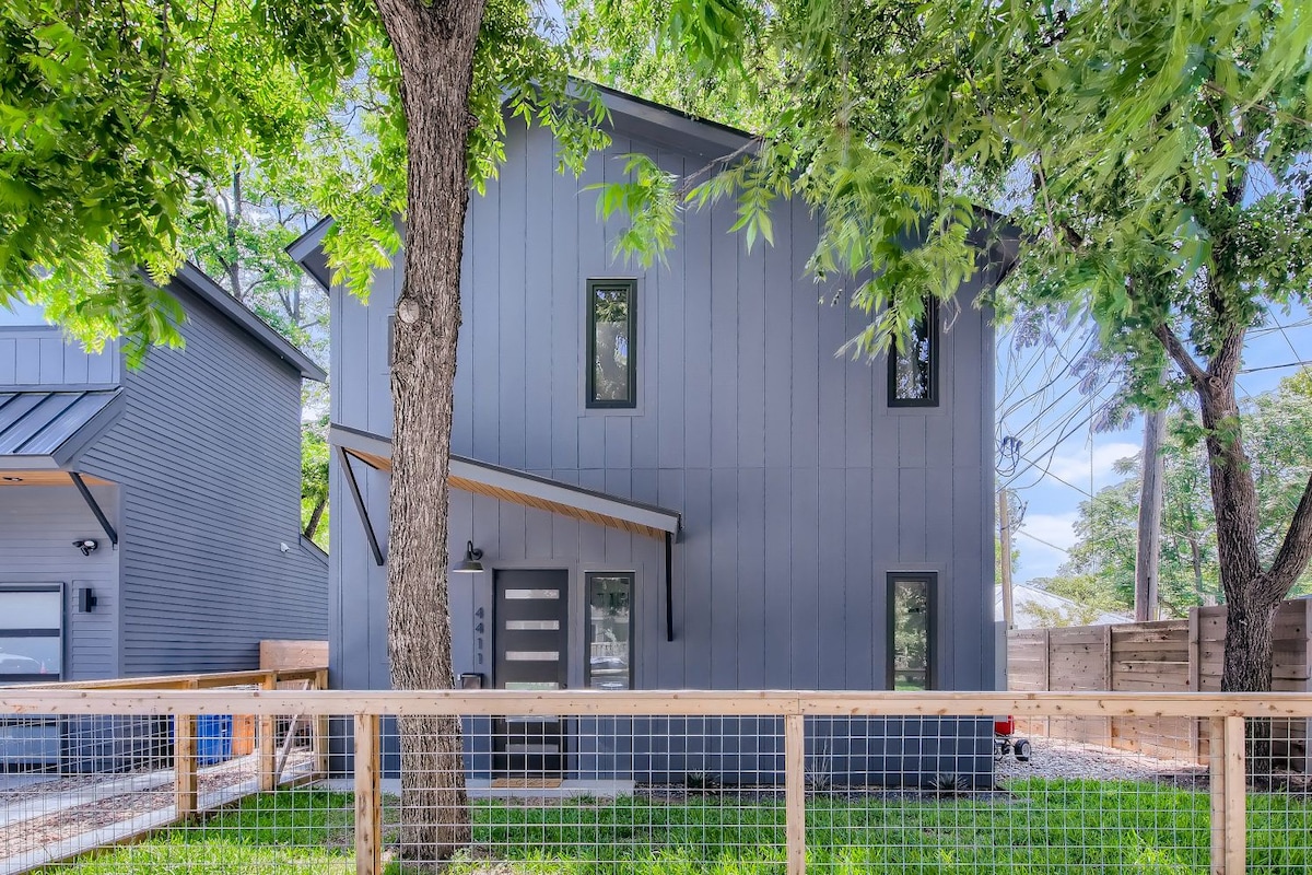 The two-story home features a modern gray exterior complemented by large windows and a covered entrance. A fenced yard with green grass surrounds the property, while mature trees provide shade, creating a welcoming environment.