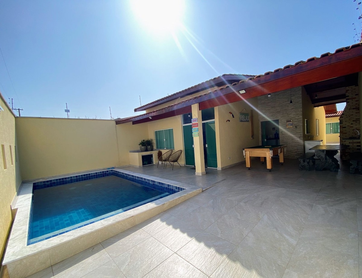 A private pool is surrounded by a spacious outdoor area, featuring light-colored tiles and a shaded seating area with chairs. A game table is positioned nearby, with the structure's wooden beams visible overhead. Sunlight streams in, creating a bright and open atmosphere.
