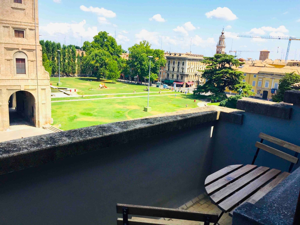 A small balcony features a round wooden table accompanied by two chairs, providing a view of a green park with trees and distant buildings. The sky is bright with scattered clouds, enhancing the open atmosphere of the setting.