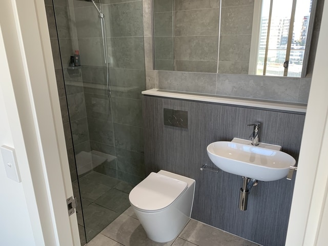 A modern bathroom is displayed, featuring a walk-in shower enclosed by glass panels and a sleek showerhead. A wall-mounted sink is positioned beside a contemporary toilet. Soft gray tiles cover the walls and floor, enhancing a clean and minimalistic aesthetic.