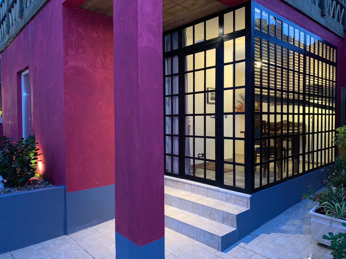 An entrance is framed by a striking maroon wall, leading to a glass-enclosed winter garden. Large black-framed glass doors provide visibility into the interior, where warm light illuminates a dining area. Steps made of neutral-toned tiles connect the outdoor space to the interior.
