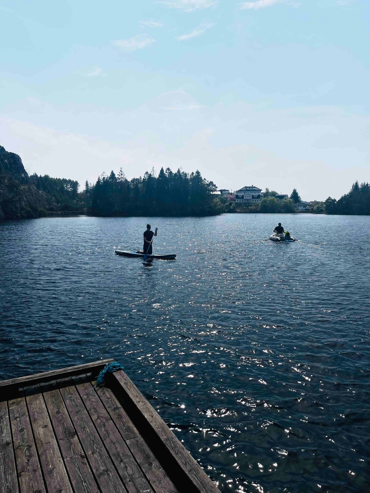 Two individuals are seen enjoying water activities on the lake. One person stands on a stand-up paddleboard while another is seated in a kayak. The still water reflects the surrounding trees and a distant building, creating a serene atmosphere.