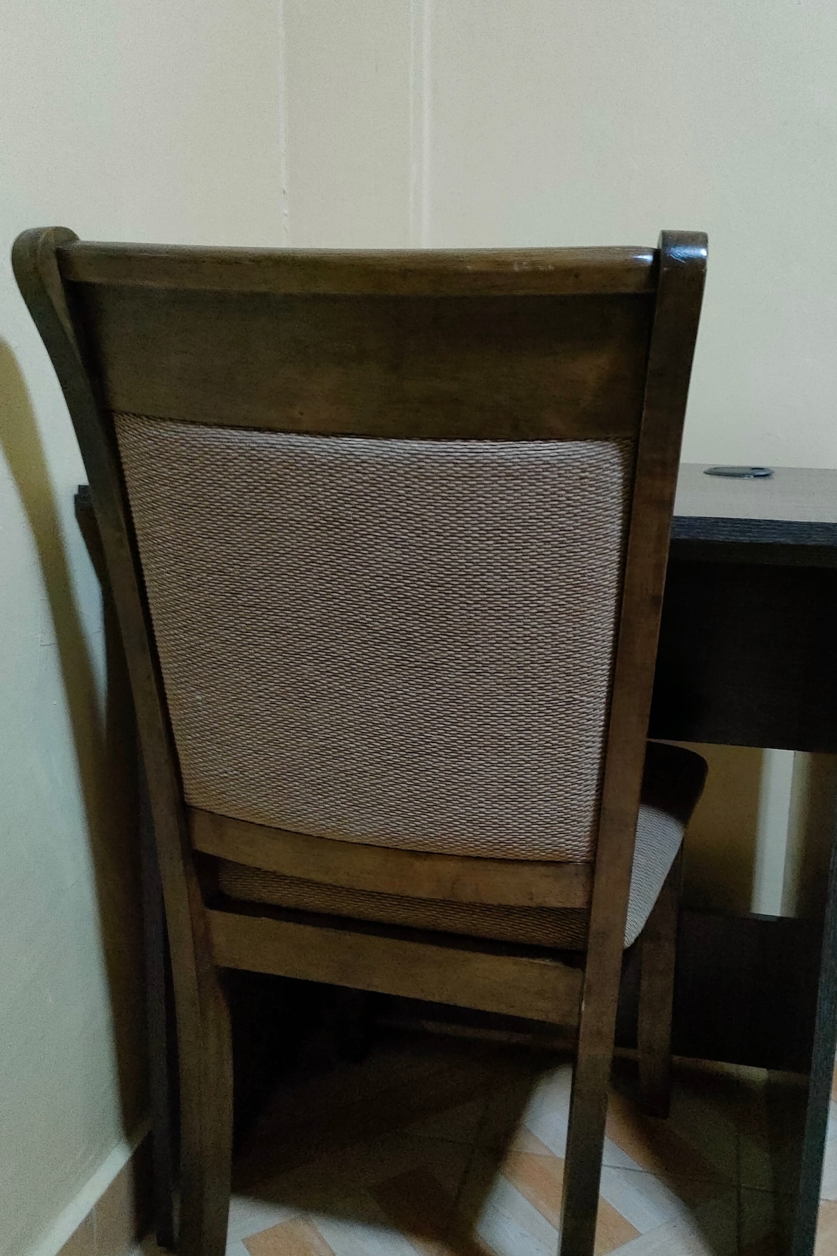 A wooden desk is shown alongside a matching chair with a woven fabric backrest. The neutral-colored wall behind them complements the furniture, providing a conducive space for work. The floor features a patterned design, adding subtle visual interest.