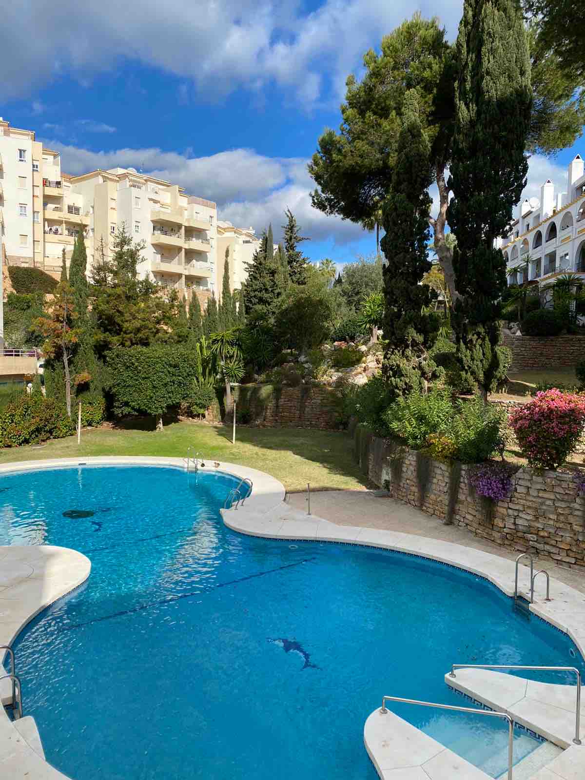 A shimmering pool is surrounded by lush greenery and flowering plants, set against a backdrop of multi-story buildings. Relaxing lounge chairs are positioned nearby, and a gently sloping lawn offers additional space for leisure. Clear blue sky adds to the inviting atmosphere.