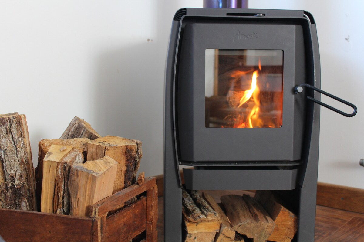 A black metal stove displays a lively fire, offering warmth. Nearby, a wooden box is filled with neatly stacked logs, providing a rustic touch. The simple design complements the cozy atmosphere of the space.