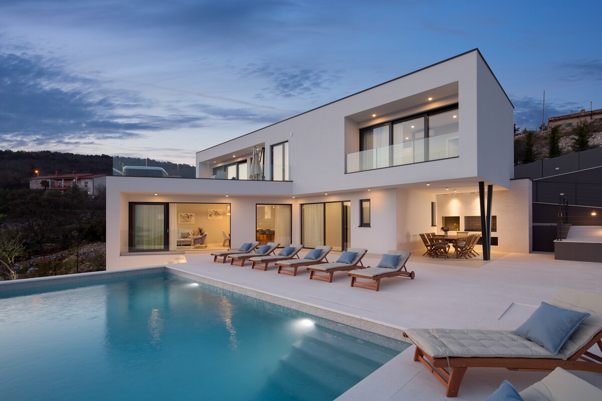 A modern villa features a spacious terrace with several lounge chairs positioned around a tranquil swimming pool. The exterior showcases clean lines and large glass doors, connecting the indoor living spaces with the outdoor setting, complemented by a backdrop of natural landscape.