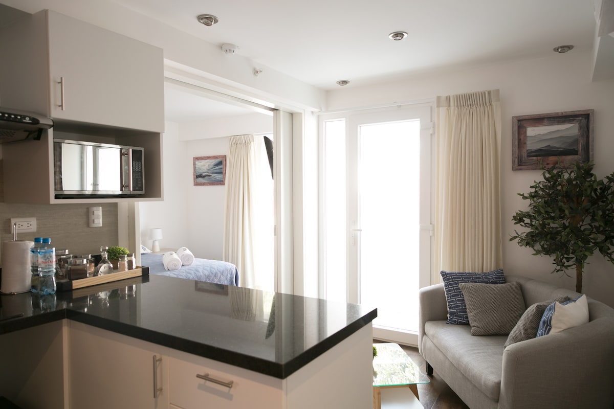 A modern kitchen area features a black countertop with a microwave and a small water bottle. The bright living space includes a comfortable gray sofa adorned with decorative pillows, and large windows allow natural light to fill the room.