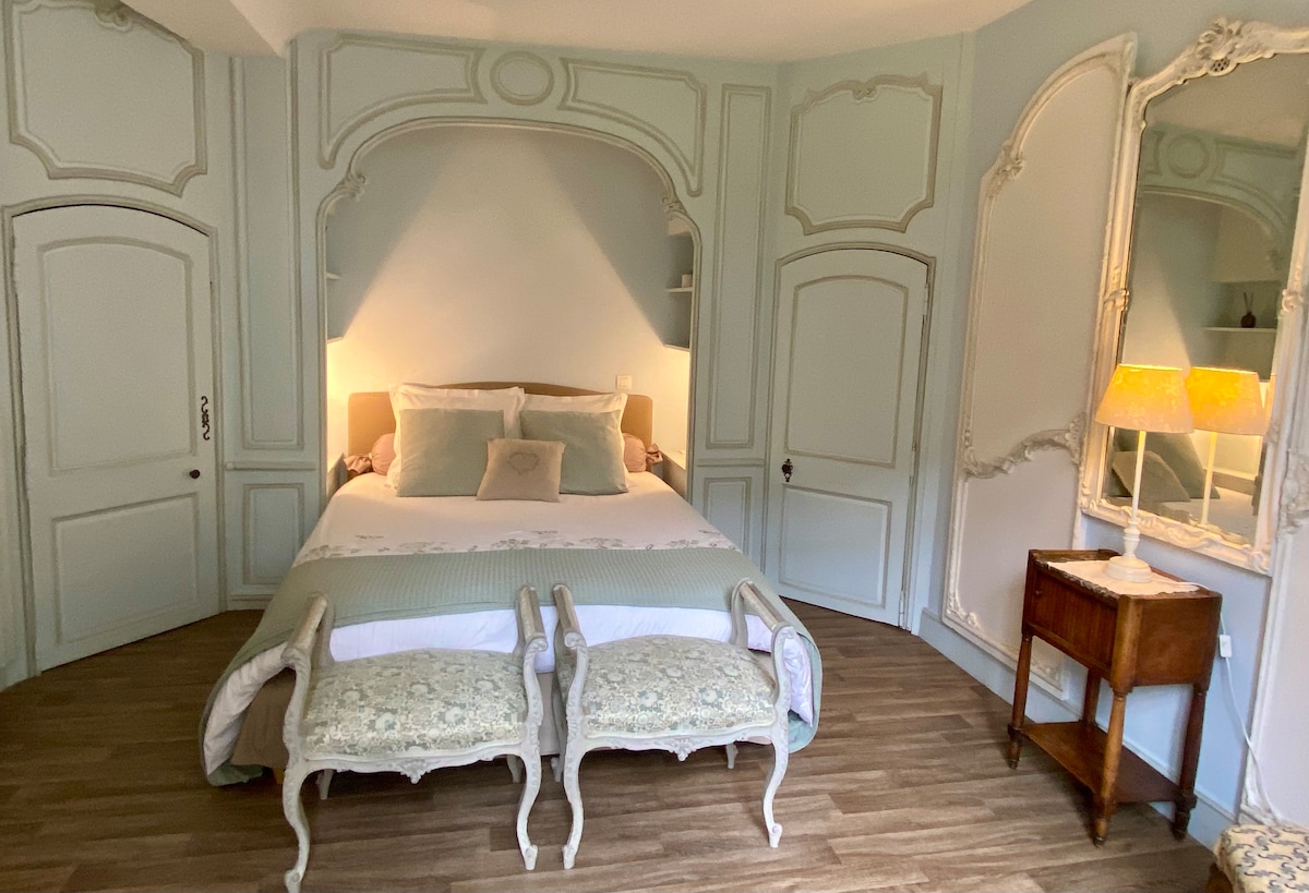 A softly lit bedroom features a spacious bed with neutral-toned bedding, framed by elegant, pale blue walls. Two decorative chairs with intricate detailing are placed at the foot of the bed, complemented by a vintage wooden nightstand with a lamp. Soft lighting adds warmth to the space.