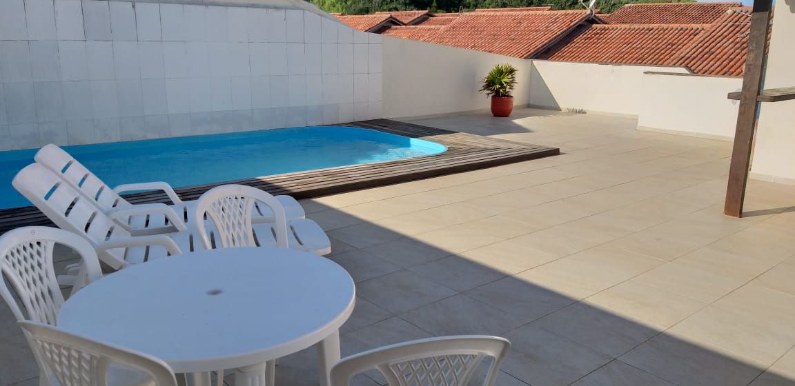 A private pool area features a turquoise pool surrounded by a white-tiled deck. Four white lounge chairs are arranged near a round table, providing space for relaxation. A potted plant adds a touch of greenery, while a wall and roof structure define the outdoor space.
