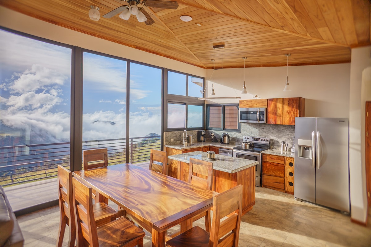 A modern kitchen and dining area is highlighted by large windows that offer sweeping views of the clouds and surrounding landscape. The space features a wooden dining table with matching chairs, stainless steel appliances, and a contrasting grey countertop.
