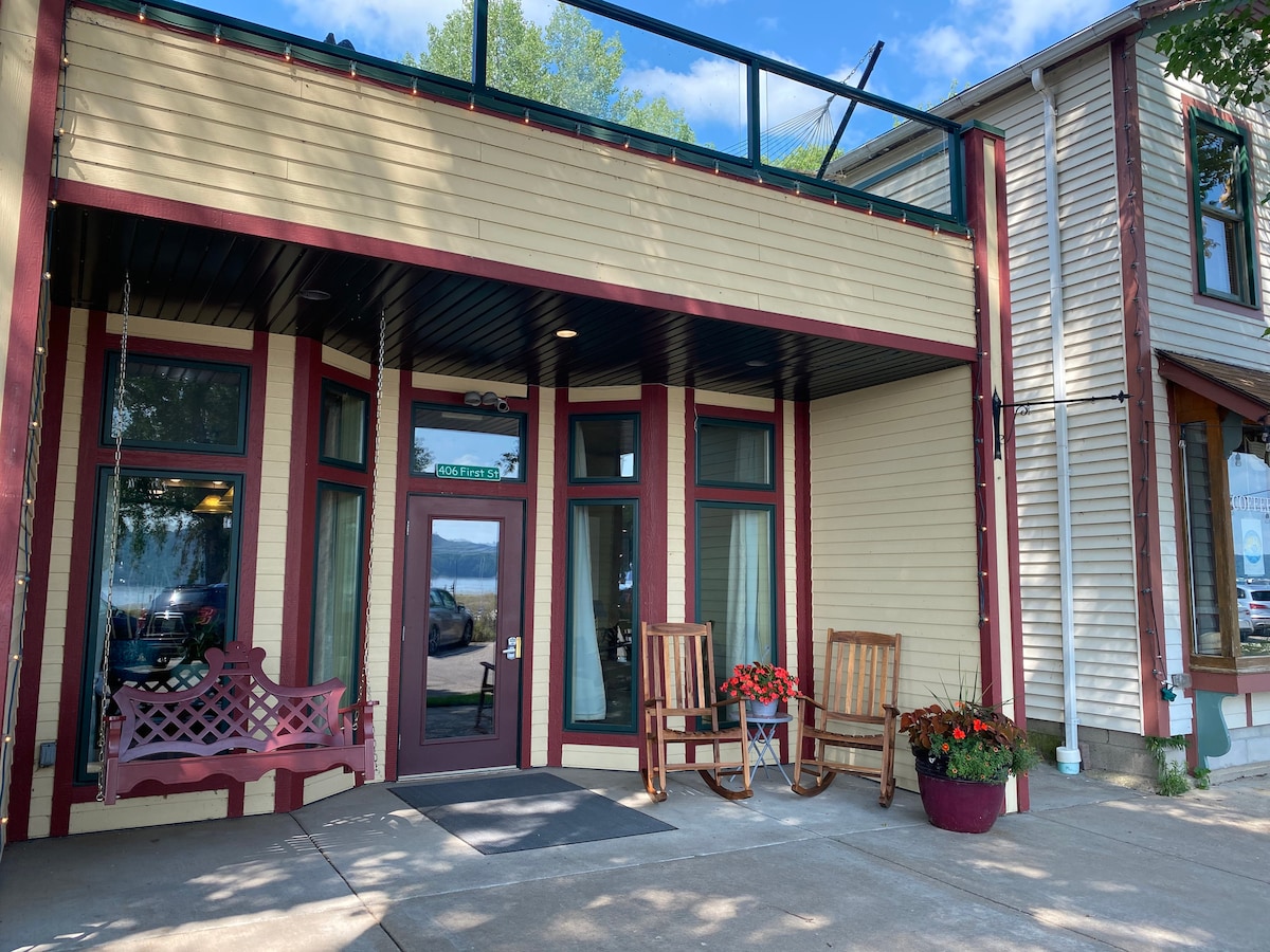 The entrance features large windows framed in dark green, complemented by two wooden rocking chairs. A vibrant flower pot adds color to the entryway, while a sign indicates the building's name. A glimpse of Lake Pepin is visible in the background.