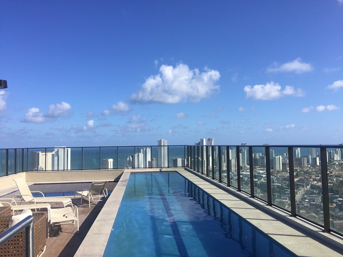 A rooftop pool is viewed, surrounded by glass railing that offers panoramic views of the city and ocean. Comfortable lounge chairs are positioned along the poolside, providing a relaxing space under a bright blue sky filled with white clouds.
