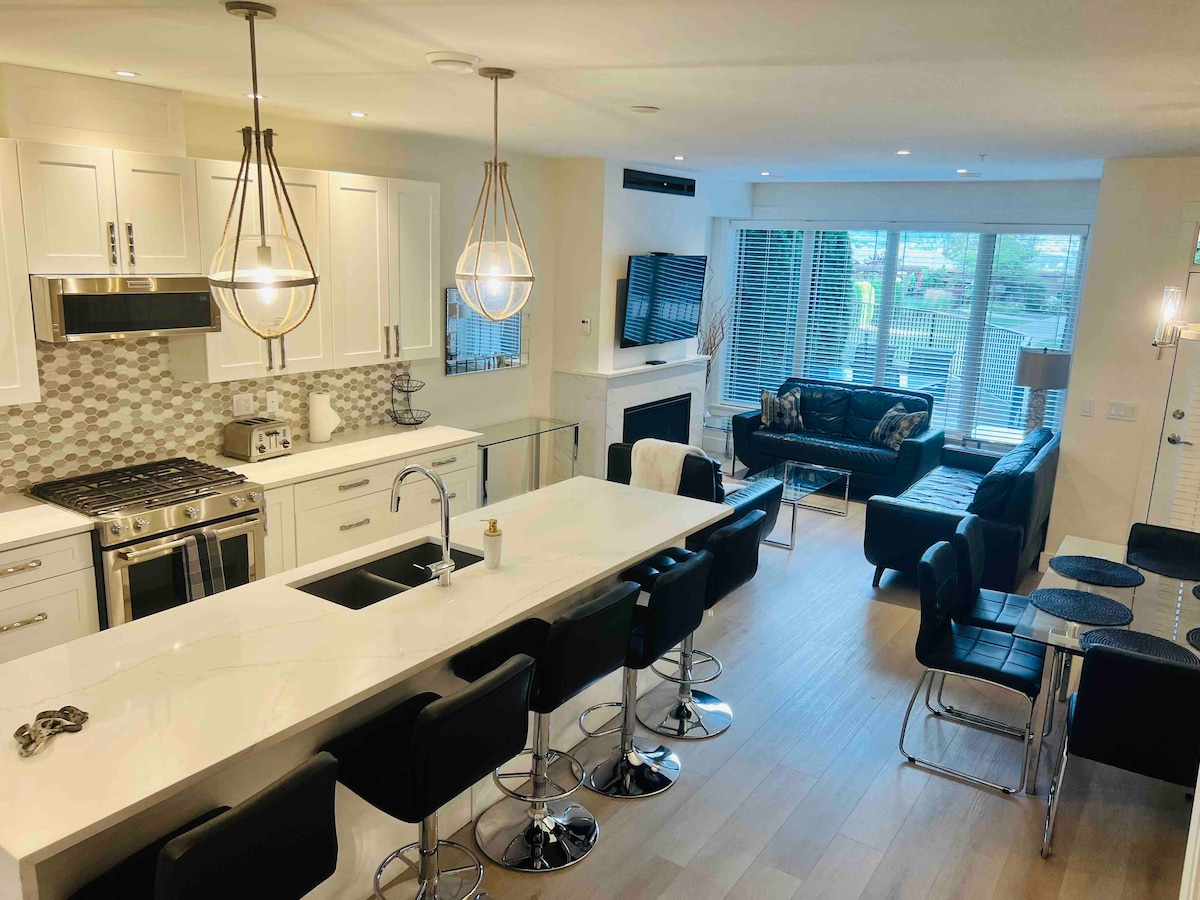 A modern kitchen area features white cabinetry, a large island with bar seating, and stainless steel appliances. The open living space is highlighted by a sleek black sofa, a flat-screen television mounted above a contemporary fireplace, and ample natural light from the large windows.