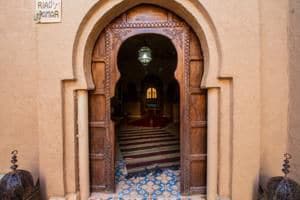 A traditional arched wooden door opens to reveal an intricate tiled entrance. Soft lighting casts a warm glow on the patterned floor, drawing attention to the ornate details of the architecture. The welcoming space beyond suggests a blend of comfort and cultural heritage.