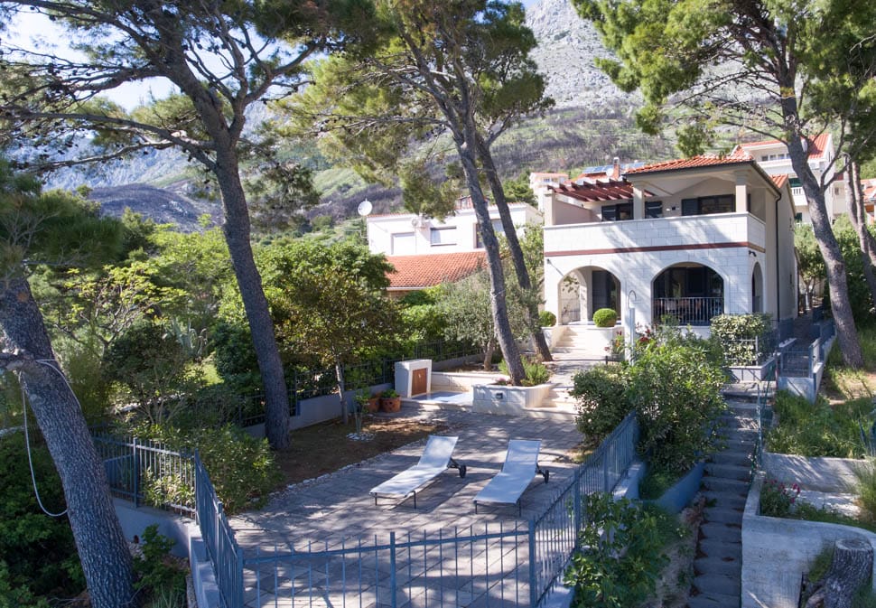 The villa is surrounded by lush greenery and towering trees, providing shade and tranquility. Lounge chairs are arranged on a stone patio, offering a place to relax. The structure features a welcoming terrace with arches, blending harmoniously with the natural landscape.