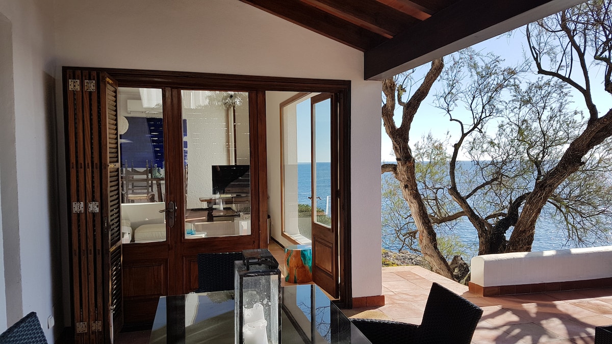 An open living area is presented, featuring large windows that provide an unobstructed view of the azure sea. A dining table is set with dark woven chairs, while a lantern sits on the table. The natural elements of the garden, including a tree, are visible through the windows.