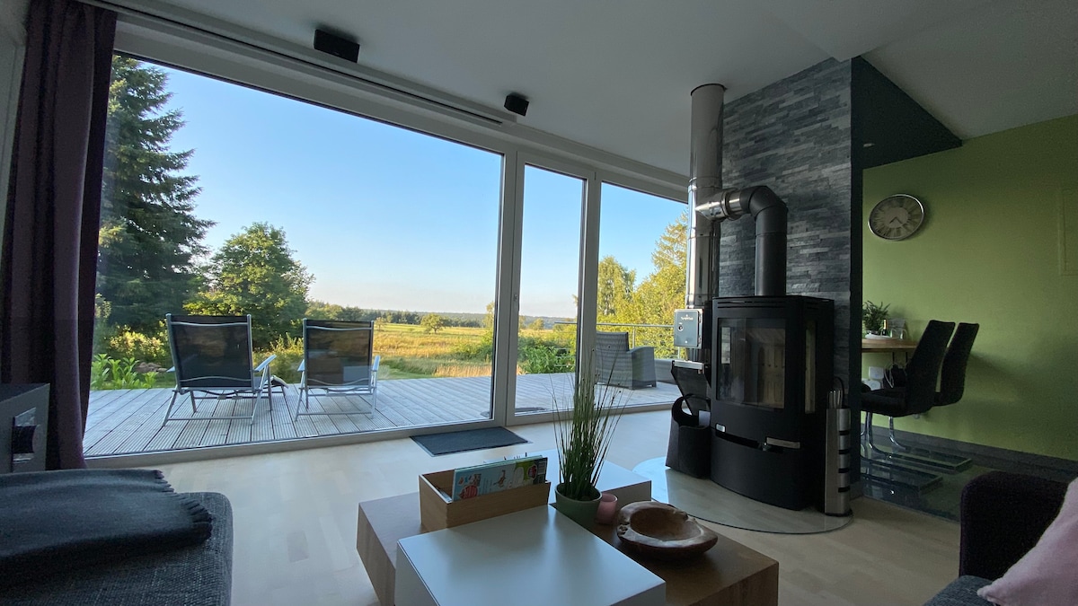 A bright living area features large glass doors revealing views of the outdoor landscape. A wood-burning stove adds warmth, and outdoor seating is positioned on the wooden terrace. Natural light fills the room, complemented by a green accent wall.