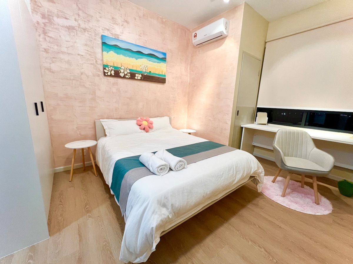 A cozy studio room features a spacious bed with soft linens and decorative pillows. Subtle pink walls complement a floral artwork above the bed. A writing desk and chair are positioned near a window, allowing natural light to illuminate the space.