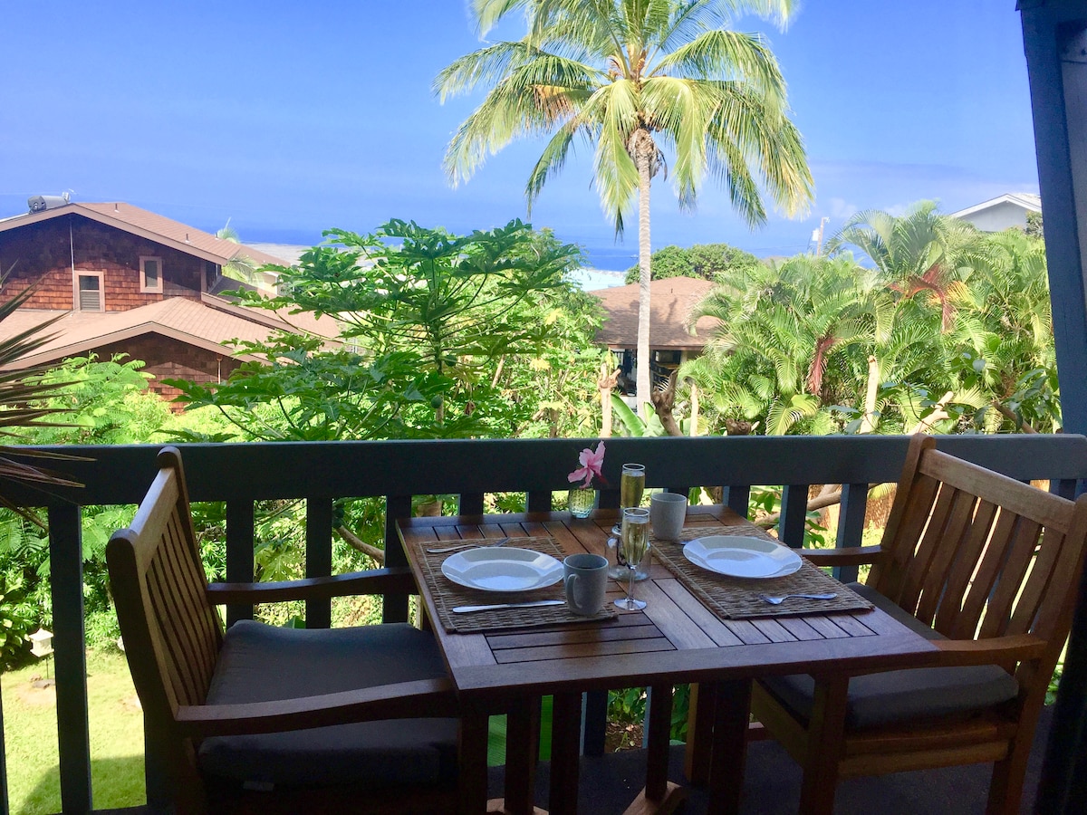 A private lanai is displayed, furnished with a wooden dining table and two chairs. The table is set with plates and mugs. Lush greenery and palm trees surround the space, offering a view of the ocean in the distance under a clear blue sky.