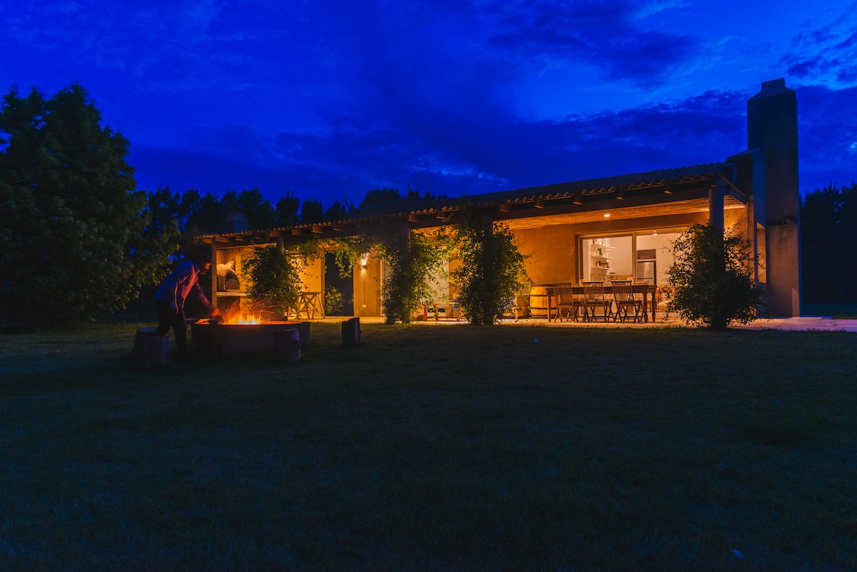 The exterior of the cottage is softly illuminated under a twilight sky, featuring a cozy fire pit surrounded by seating. Warm light spills from the windows, highlighting the inviting open space that connects to the surrounding green pastures.