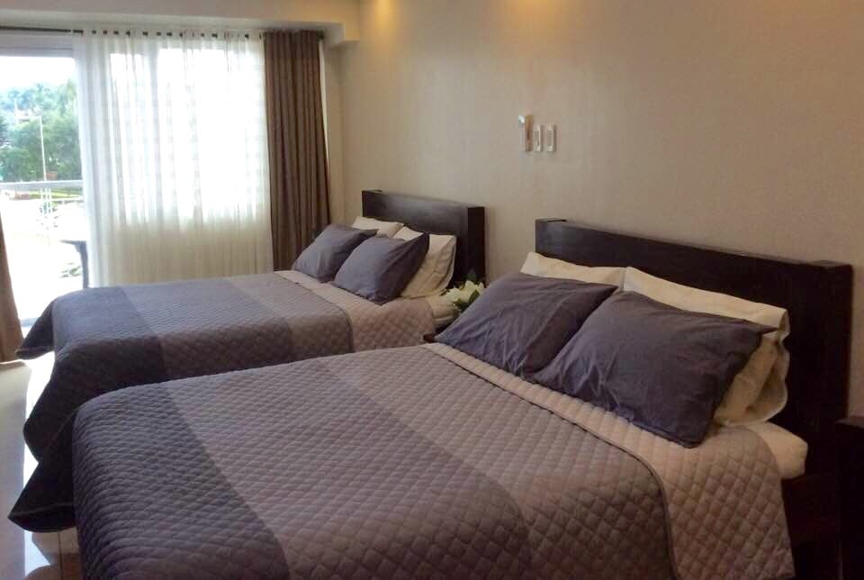 The bedroom features two queen-sized beds, each adorned with simple bed linens and accent pillows. Natural light floods the space through large windows, complemented by modern curtains. A light-colored wall adds to the room's brightness, creating an airy and spacious ambiance.