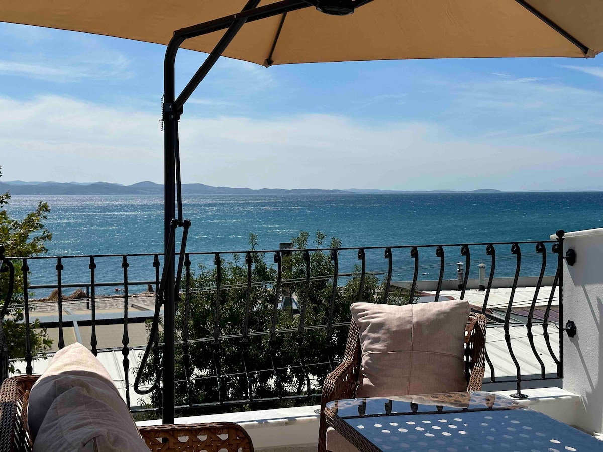 An outdoor seating area is presented with comfortable wicker chairs and a glass table. An umbrella provides shade while offering views of the glistening sea and distant hills under a clear sky.