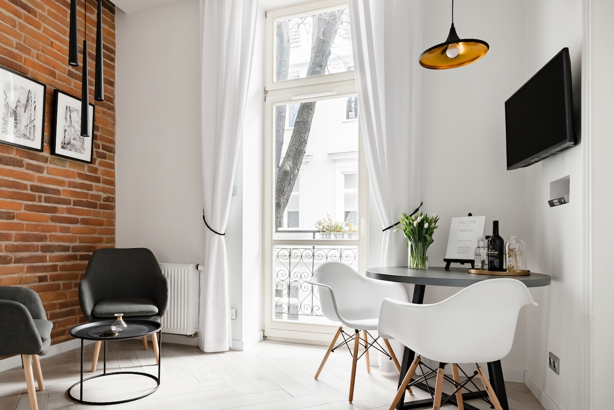A modern dining area is showcased with a round table surrounded by white chairs. A sleek black table sits beside the chairs. An exposed brick wall adds character, while large windows allow natural light to fill the space, framed by white curtains.