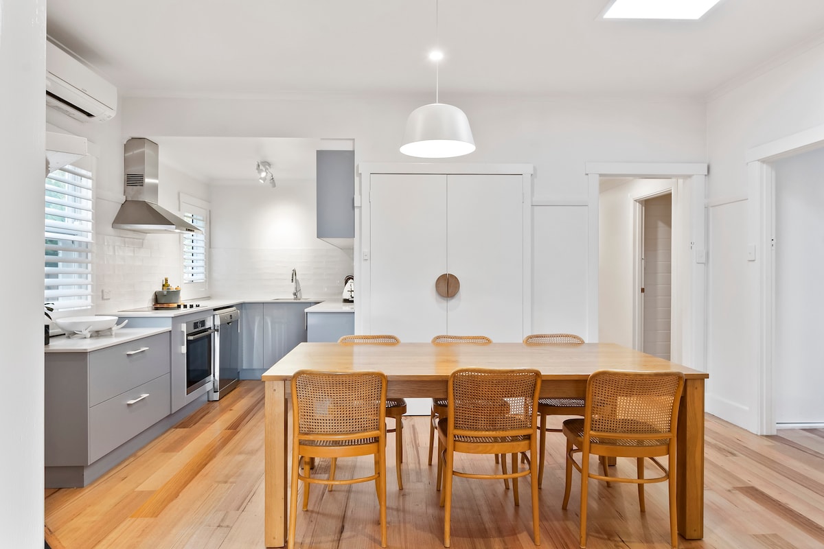 The kitchen and dining area features light wood flooring and a large wooden dining table surrounded by six rattan chairs. Modern appliances are present in the kitchen, along with sleek cabinetry. Natural light enters through a skylight and window, enhancing the open layout.