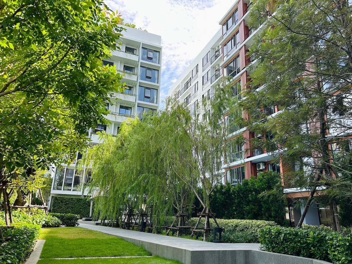A landscaped courtyard is framed by modern apartment buildings, featuring lush greenery and tall trees. A pathway leads through the grassy area, providing a serene environment for relaxation. The design harmoniously blends nature with contemporary architecture.