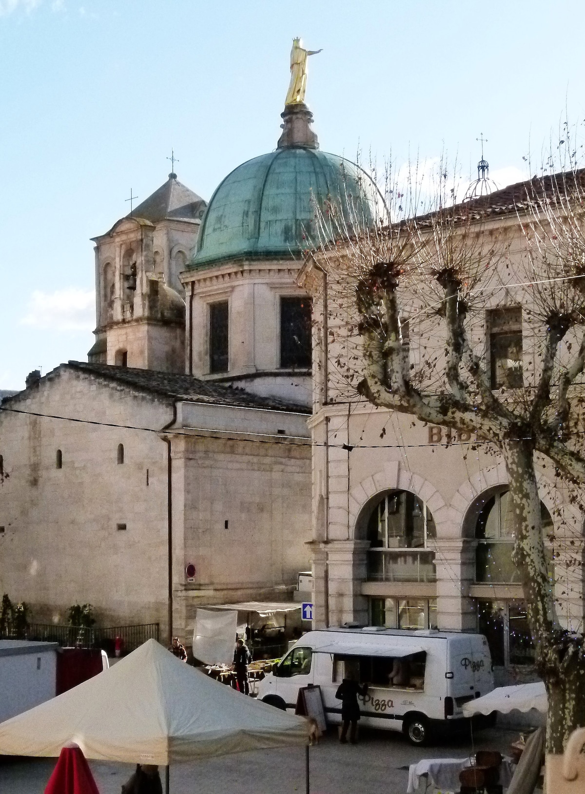 A scenic view features a historic building with a distinctive dome topped by a statue. The structure has a muted stone façade, complemented by a tree with bare branches nearby. A lively square is visible below, where a food truck and market stalls are set up.