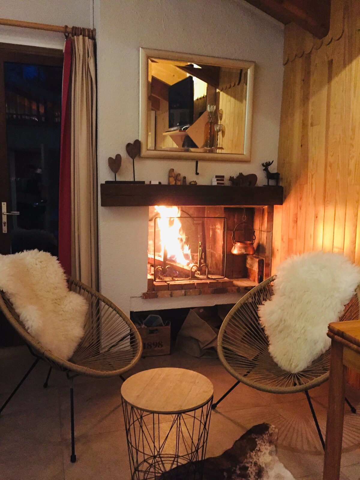 A cozy seating area features two woven chairs adorned with soft white throws, positioned around a fireplace. The flame is visible, providing warmth and illumination, while a small wooden table sits nearby. A rustic mirror and wooden accents enhance the welcoming ambiance.