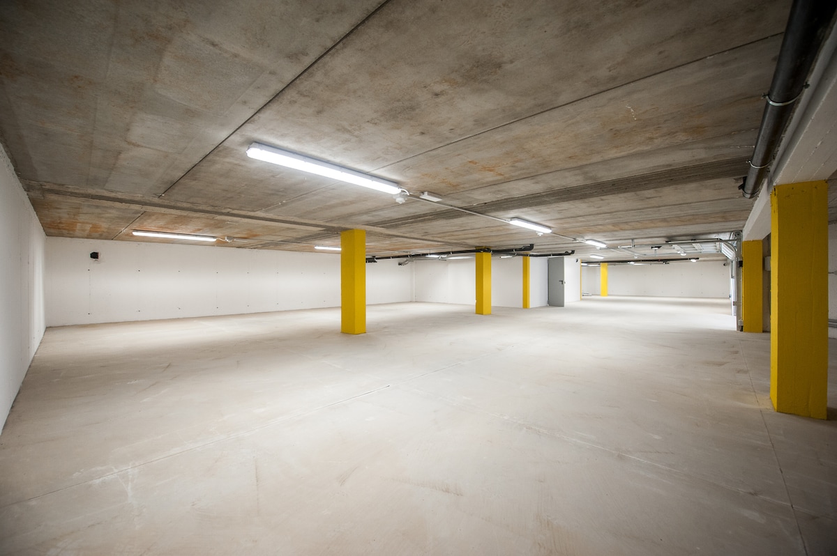 A spacious underground garage is depicted, showcasing expansive concrete flooring and bright overhead lighting. Yellow pillars provide support throughout the area, and a clear view of the ample space is presented. The walls are unadorned and painted white, contributing to a clean and modern appearance.