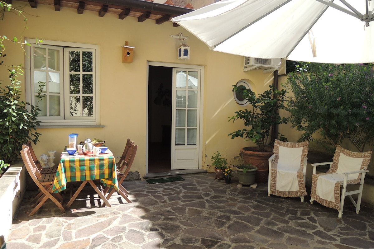 A sunlit courtyard is depicted, featuring a stone-paved floor. A dining table draped with a checkered cloth is set for a meal, surrounded by wooden chairs. Two lounge chairs, positioned nearby, offer a comfortable space for relaxation. The entrance to the cottage is visible, framed by green plants.