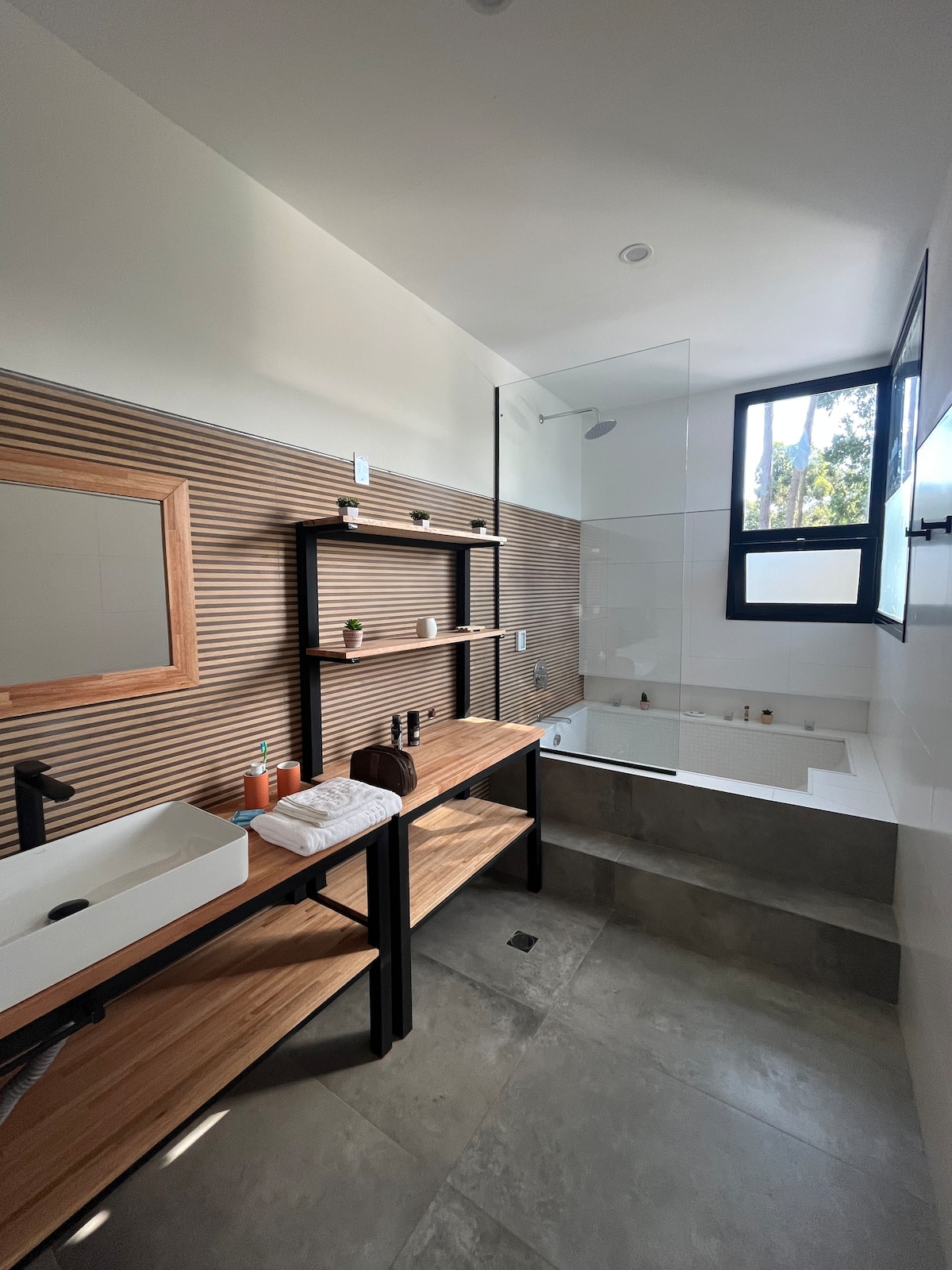 The spacious bathroom features wood-paneled walls and modern fixtures. A sleek glass shower is positioned beside a sunken bathtub. Open shelving displays toiletries, while a large mirror is mounted above a stylish sink. Natural light filters in through a nearby window.
