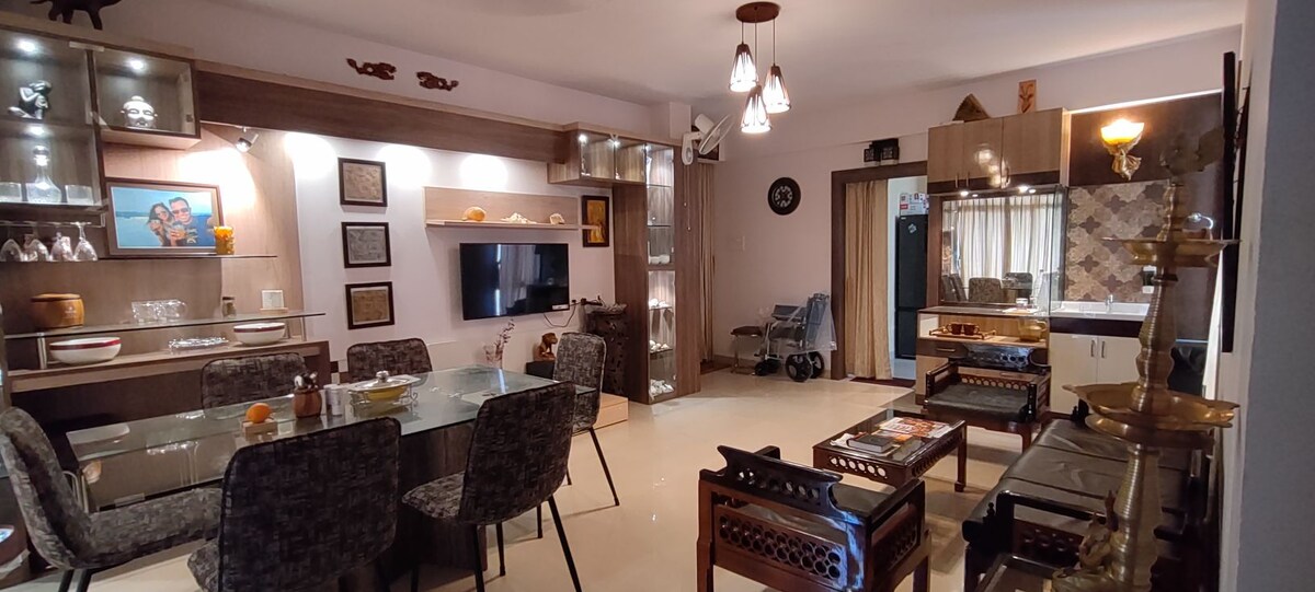 A modern dining area features a glass table surrounded by patterned chairs. A wall-mounted television is visible, along with decorative items on open shelves. Soft lighting emanates from stylish fixtures, creating a welcoming environment. A glimpse of the kitchen area is presented in the background.